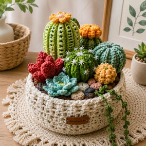 Op de afbeelding: Een verzameling gehaakte cactussen en vetplanten in een ronde, crèmekleurige gehaakte mand. De planten hebben verschillende tinten groen, rood, geel en blauw garen, met kleine witte stervormige accenten. Een bruin leren label met de tekst Hand made is bevestigd aan de voorzijde van de mand.