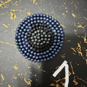 May include: A circular brooch featuring concentric rings of small, metallic blue beads surrounding a central cluster of dark, faceted black beads. The design is symmetrical and compact, with the number one written in white below the object.