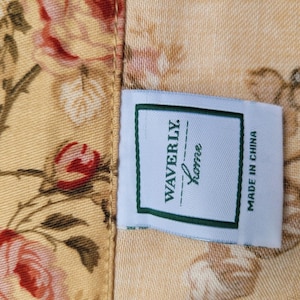 May include: A fabric tag with a white background and a green border featuring the text WAVERLY Home and MADE IN CHINA, attached to a yellow textile surface decorated with a floral pattern of pink roses and green leaves on brown stems.