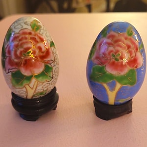 May include: Two egg-shaped ornaments featuring floral designs with pink flowers and green leaves. One egg has a white background with a cloud pattern, while the other has a solid blue background. Both are displayed on small, round, black stands.