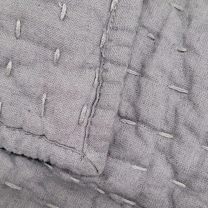May include: A close-up view of a grey textile featuring a quilted texture with visible running stitches in a lighter grey thread. The fabric has a soft, slightly wrinkled appearance with a distinct hemmed edge showing layered construction.
