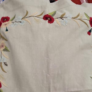 May include: A light beige fabric featuring an embroidered floral border. The design includes red poppies, light blue flowers, and green leaves arranged in a curved pattern along the edges of the textile, showcasing detailed needlework.