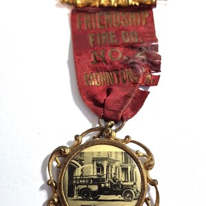 May include: A vintage gold-tone metal badge featuring a rectangular top bar with the word MEMBER, a red fabric ribbon printed with FRIENDSHIP FIRE CO. NO. 2 MOHNTON PA, and a circular pendant displaying a black and white photograph of a fire truck.