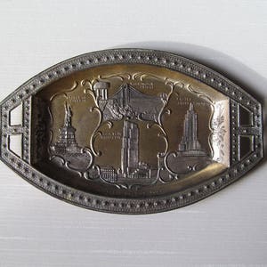 Puede incluir: Bandeja ovalada de metal con centro dorado y borde plateado. La bandeja presenta im&aacute;genes en relieve de monumentos de la ciudad de Nueva York, como la Estatua de la Libertad, un puente y el Empire State Building.