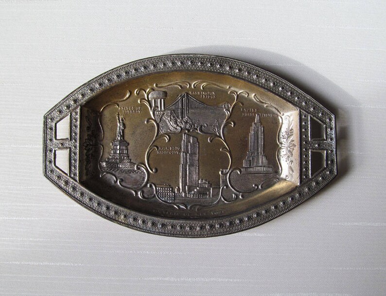 Puede incluir: Bandeja ovalada de metal con centro dorado y borde plateado. La bandeja presenta im&aacute;genes en relieve de monumentos de la ciudad de Nueva York, como la Estatua de la Libertad, un puente y el Empire State Building.
