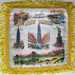 May include: White souvenir pillow with a yellow fringe, featuring colorful illustrations of New York City landmarks, including Times Square, the Empire State Building, and the Statue of Liberty. The text "Souvenir of New York" is displayed.