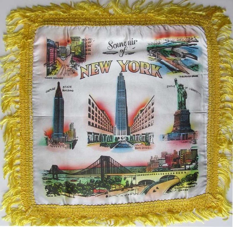 May include: White souvenir pillow with a yellow fringe, featuring colorful illustrations of New York City landmarks, including Times Square, the Empire State Building, and the Statue of Liberty. The text "Souvenir of New York" is displayed.