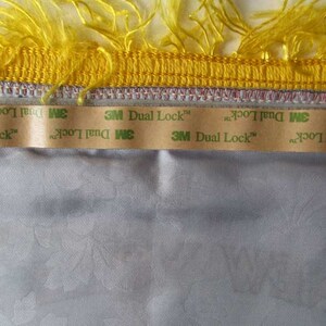 May include: Close-up of a decorative trim with yellow fringe and a gold border. Below the trim is a strip of 3M Dual Lock tape, adhered to a light gray fabric with a subtle floral pattern. The image highlights the texture and details.