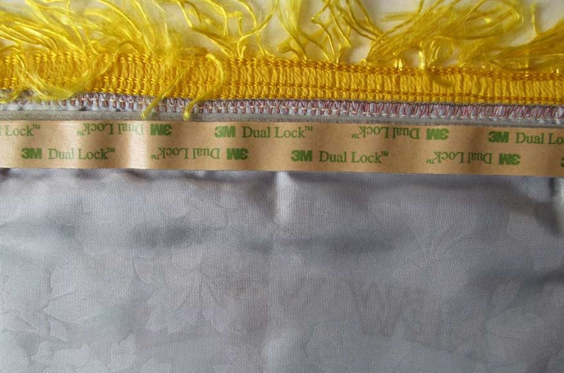 May include: Close-up of a decorative trim with yellow fringe and a gold border. Below the trim is a strip of 3M Dual Lock tape, adhered to a light gray fabric with a subtle floral pattern. The image highlights the texture and details.
