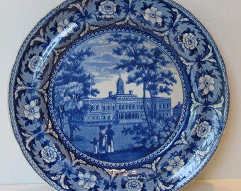 May include: A round, blue and white decorative plate. The plate features a central landscape scene with a building and figures, surrounded by a floral and leaf border. The plate's design is a classic, vintage style.