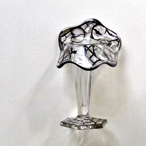 May include: A clear glass bud vase with a flared, scalloped top and a geometric base. The top and base feature a black, web-like design. The vase has a slender stem and is suitable for displaying a single flower or small bouquet.