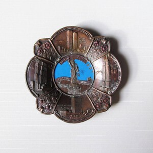 Puede incluir: Un plato de recuerdo de metal en tono bronce con un dise&ntilde;o floral. El centro presenta un c&iacute;rculo azul con la Estatua de la Libertad. El plato tambi&eacute;n representa las Torres Gemelas, el Empire State Building y la sede de las Naciones Unidas.