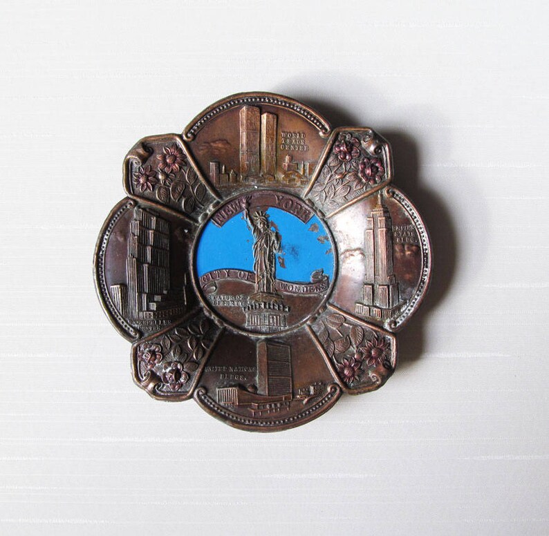 Puede incluir: Un plato de recuerdo de metal en tono bronce con un dise&ntilde;o floral. El centro presenta un c&iacute;rculo azul con la Estatua de la Libertad. El plato tambi&eacute;n representa las Torres Gemelas, el Empire State Building y la sede de las Naciones Unidas.