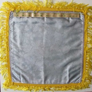 May include: A square, light blue satin pillow cover with a floral pattern. The pillow has a yellow fringe border and a silver trim. The text "3M Dual Lock" is visible on the top edge of the pillow.