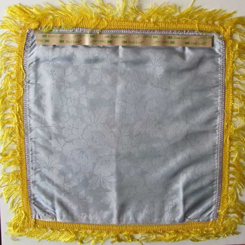 May include: A square, light blue satin pillow cover with a floral pattern. The pillow has a yellow fringe border and a silver trim. The text "3M Dual Lock" is visible on the top edge of the pillow.