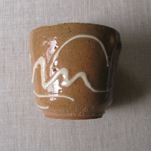May include: A small, handmade ceramic cup with a brown exterior and a white, wavy design. The cup has a slightly textured surface and a reddish-brown base. The design resembles a stylized mountain range.