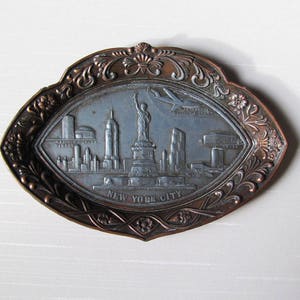 Puede incluir: Bandeja met&aacute;lica de color bronce, de forma ovalada, con un dise&ntilde;o en relieve del horizonte de Nueva York. La bandeja presenta la Estatua de la Libertad, rascacielos y la inscripci&oacute;n "NEW YORK CITY". El borde tiene un patr&oacute;n floral ornamentado.
