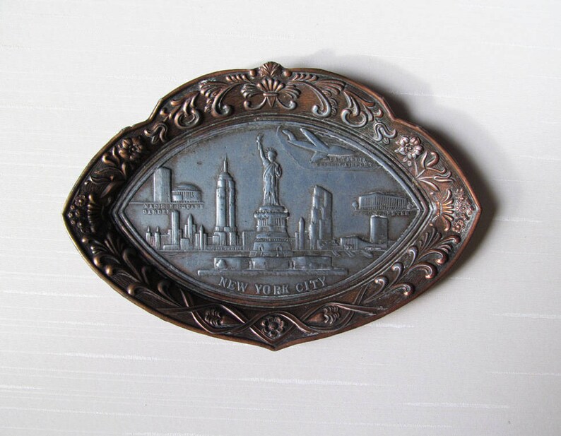Puede incluir: Bandeja met&aacute;lica de color bronce, de forma ovalada, con un dise&ntilde;o en relieve del horizonte de Nueva York. La bandeja presenta la Estatua de la Libertad, rascacielos y la inscripci&oacute;n "NEW YORK CITY". El borde tiene un patr&oacute;n floral ornamentado.