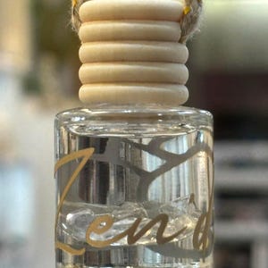 Puede incluir: Un frasco pequeño de vidrio transparente lleno de líquido y cristales pequeños, con la palabra Zen en letras doradas. El frasco tiene una tapa de madera clara acanalada y está suspendido por una cuerda gruesa, trenzada y beige con hilo dorado.