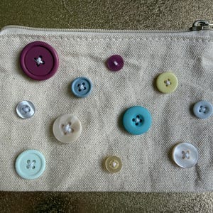 May include: A rectangular beige canvas zippered pouch decorated with eleven assorted buttons in various sizes and colors, including shades of purple, blue, teal, yellow, and white, each securely stitched onto the front fabric surface.