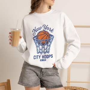 May include: A white long-sleeve sweatshirt featuring a graphic print of a basketball entering a net. The text reads New York CITY HOOPS EST. 1978 in blue lettering. The design is centered on the chest of the garment, which is paired with gray denim shorts.