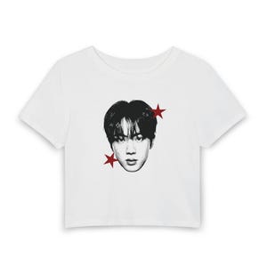 Op de afbeelding: Een wit kort T-shirt met korte mouwen en een zwart-wit grafisch portret van een persoon met donker haar in het midden op de borst, geflankeerd door twee kleine, rode, vijfpuntige sterren rechtsboven en linksonder van het gezicht.