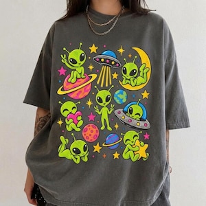 May include: A charcoal grey short-sleeve t-shirt featuring a colourful graphic print of multiple green aliens, flying saucers, planets, and stars. The design includes various cartoon-style extraterrestrial figures interacting with space elements.