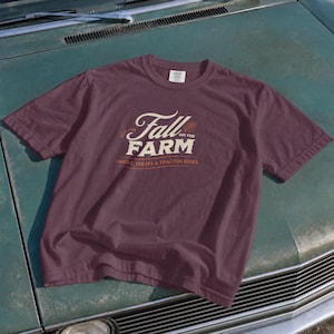 May include: A dark maroon short-sleeve t-shirt featuring the cream-colored text Fall on the Farm, with the smaller orange text Tricks, Treats & Tractor Rides below. A small leaf icon is positioned to the right of the word Fall on the chest.