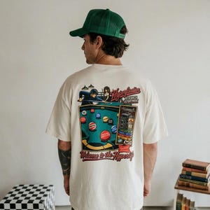 May include: An off-white short-sleeve t-shirt featuring a colorful graphic on the back. The design depicts a pool table with billiard balls, a person playing pool, a vintage camera, and the text "Hyperfixation Research Department" and "Welcome to the Hyperlife."