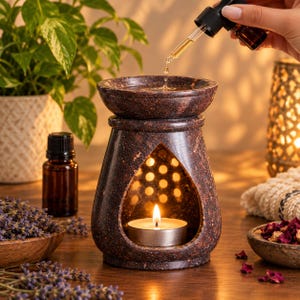 Puede incluir: Un quemador de aceite de piedra moteada de color marrón oscuro con un cuenco ancho en la parte superior y una abertura en forma de lágrima en el frente. Una vela de té encendida descansa en la base mientras un gotero vierte aceite en el cuenco.