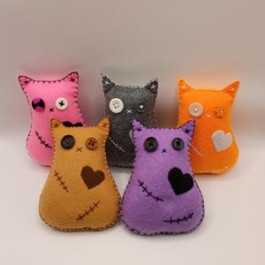 May include: Five small, plush cat-shaped dolls made of felt in pink, gray, orange, tan, and purple. Each doll features button eyes, stitched mouth details, and decorative felt patches in heart or flower shapes, with visible whip-stitch edging.
