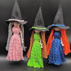 May include: Three hanging witch ornaments made of yarn and felt. Each features a black pointed hat with a colored band, a round wooden bead head, and long yarn hair. The dolls wear dresses in pink, bright green, and blue with matching yarn hair.