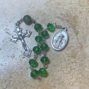 Puede incluir: Un rosario con cuentas facetadas verdes y un crucifijo plateado. El rosario también presenta un colgante plateado con la imagen de un santo. Las cuentas están ensartadas en una cadena plateada.