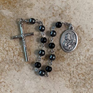 Op de afbeelding: Een rozenkrans met een zilveren kruisbeeld en een hanger met een religieuze figuur. De rozenkrans heeft afwisselend zwarte en grijze kralen. Het kruisbeeld toont een gedetailleerde afbeelding van Jezus. De hanger is ovaal van vorm.