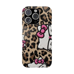 Peut inclure: Une coque de smartphone avec un motif imprimé léopard beige et noir, ornée de plusieurs visages de chat blancs brodés. Chaque personnage porte un nœud rose vif, des moustaches noires et a les yeux fermés, créant un design texturé.