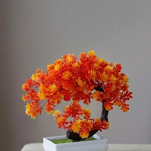 Peut inclure: Un petit bonsaï artificiel aux feuilles orange et jaune vif disposées en une canopée arrondie. L'arbre présente un tronc incurvé brun foncé et est planté dans un récipient rectangulaire en céramique blanche avec quatre petits pieds.