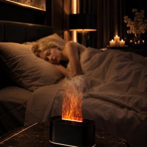Può includere: Un diffusore elettronico nero di forma ovale con un effetto fiamma simulato che emette una calda luce arancione e una sottile nebbia dalla parte superiore, con una discreta striscia luminosa alla base del dispositivo.