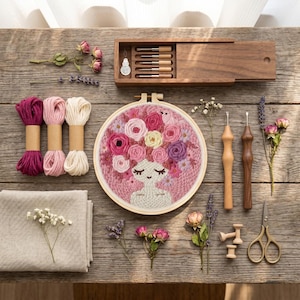 Könnte beinhalten: Ein Stickrahmen mit einem gestickten Porträt einer Figur mit Blumenkranz, begleitet von drei Garnsträngen, zwei Holz-Punch-Nadeln, einer Holzaufbewahrungsbox mit Metallspitzen, einer Schere und getrockneten Blumen auf einer rustikalen Holzoberfläche.