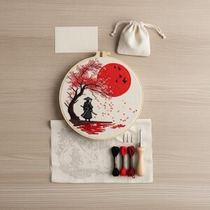 Peut inclure: Un kit de broderie comprenant un tambour en bois avec une scène brodée représentant une silhouette au chapeau conique sous un cerisier en fleurs et un grand soleil rouge. Inclut un tissu avec motif imprimé, trois échevettes de fil noir et rouge, un outil à aiguille à manche en bois et une petite pochette à cordon.
