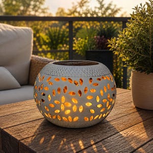 May include: A spherical white lantern with a textured surface featuring numerous leaf-shaped cutouts that emit a warm, glowing light from within. The top of the lantern has a flat, circular panel, and the overall design is rounded and compact.