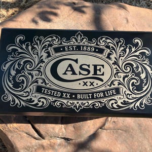 May include: A rectangular black metal storage box featuring a decorative cream-coloured filigree border. The centre displays the text CASE XX, EST. 1889, and the phrase TESTED XX BUILT FOR LIFE, all printed in a bold, stylised font on the lid.
