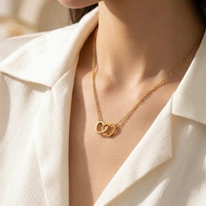 Puede incluir: Un collar de tono dorado que presenta dos colgantes entrelazados en forma de corazón suspendidos de una cadena delicada. El metal tiene un acabado liso y pulido, y el diseño consiste en dos corazones de marco abierto unidos en el centro.