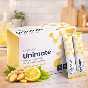 May include: A white rectangular box of Unicity Unimate ultra-concentrated yerba mate drink mix in natural lemon flavor, accompanied by two matching single-serve packets, fresh ginger root slices, and a lemon slice on a wooden surface.