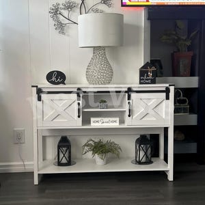 May include: A white wooden console table with two sliding barn-style doors featuring an X-pattern, black metal hardware, and an open center shelf. The table displays a white lamp, a black sign reading hi, and a black sign reading welcome HOME.