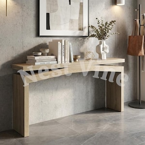 May include: A light oak console table with a unique overlapping top design. The table surface holds a stack of books titled Wabi-Sabi, Architectural Digest, and Bauhaus, alongside a white ceramic vase with eucalyptus branches and a gold geometric object.