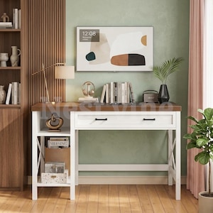 May include: A white desk with a dark wood top featuring a single drawer with two black handles, an open side shelf unit with X-shaped side supports, and a lower storage area. The desk is positioned against a sage green wall with a framed abstract art piece.