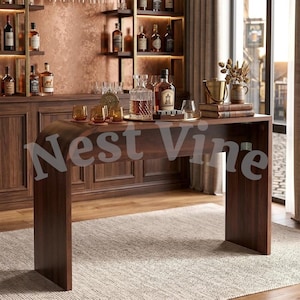 May include: A dark wood console table with a curved waterfall edge design. The surface holds a glass decanter filled with amber liquid, a bottle of liquor, three small amber glasses, a metal ice bucket, and two stacked books on a tray.