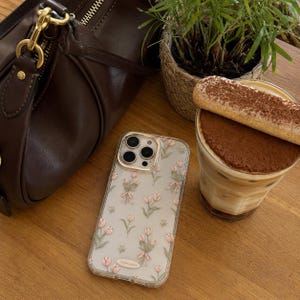 May include: A clear smartphone case featuring a repeating pattern of small pink tulips with green stems and light pink bows. The case has a gold-toned camera lens frame and a small rectangular label at the bottom with the text Tulip pattern.