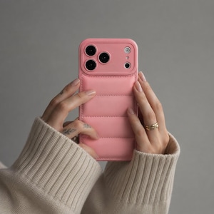 May include: A light pink smartphone case with a quilted, puffer-style texture on the back. The case features a raised rectangular camera cutout area with three circular lens openings and a small circular flash opening in the upper right corner.