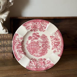 May include: A round ceramic plate featuring a red and white transferware pattern. The center depicts a landscape with a house, trees, and a bridge, while the rim is decorated with floral motifs and a dotted border on a cream-colored background.